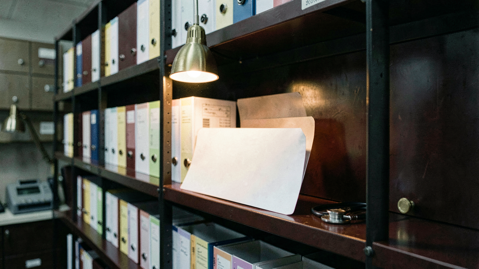 A steel shelving wall of blank colour-tabbed patient folders, one pulled out under a lamp, a stethoscope coiled beside it.